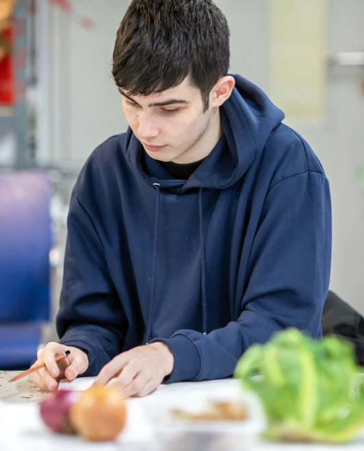 Student in a navy hoodie sketching or working on an art piece, with various fruits and vegetables on the table. Student in a navy hoodie sketching or working on an art piece, with various fruits and vegetables on the table.